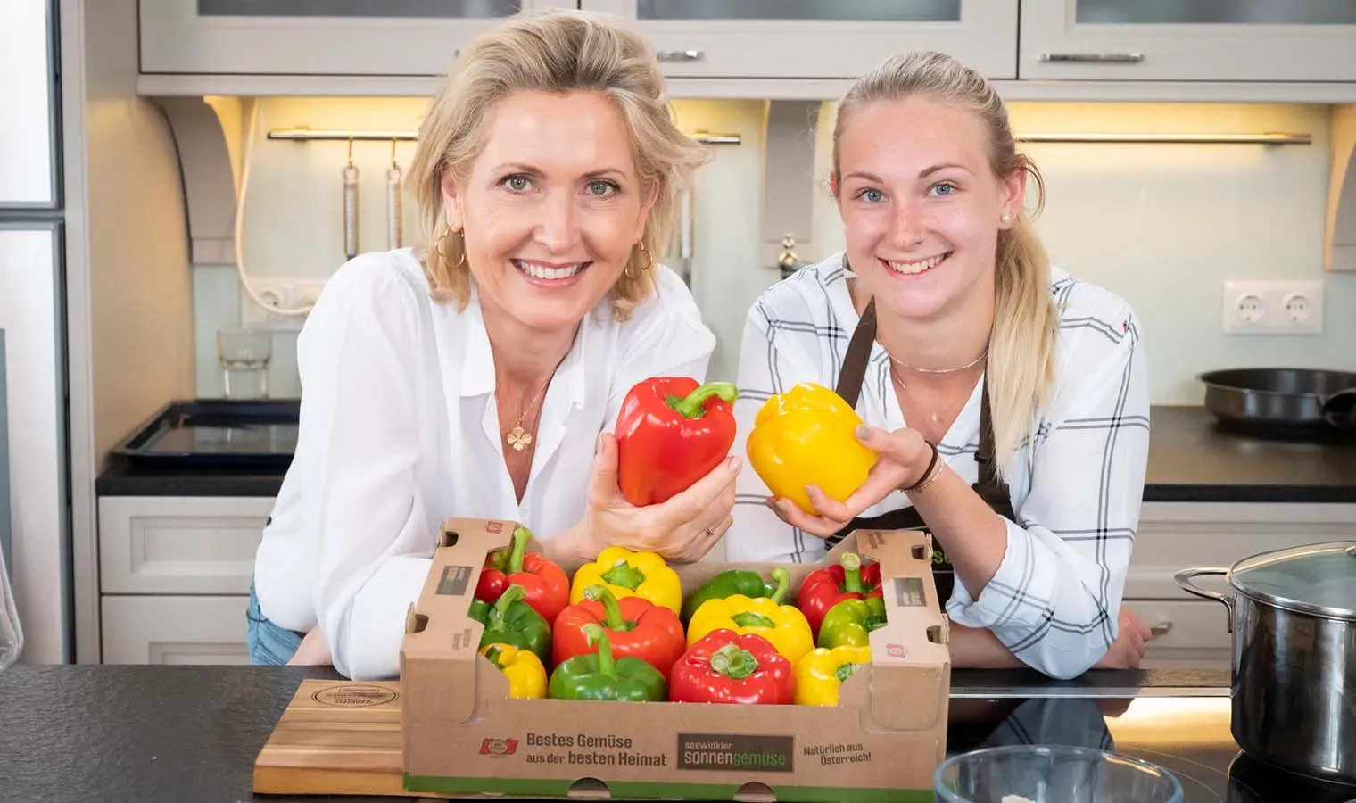 Martina Hohenlohe und Lisa Thell zeigen in der Küche rote und gelbe Paprika (Seewinkler Sonnengemüse) aus einer Gemüsekiste