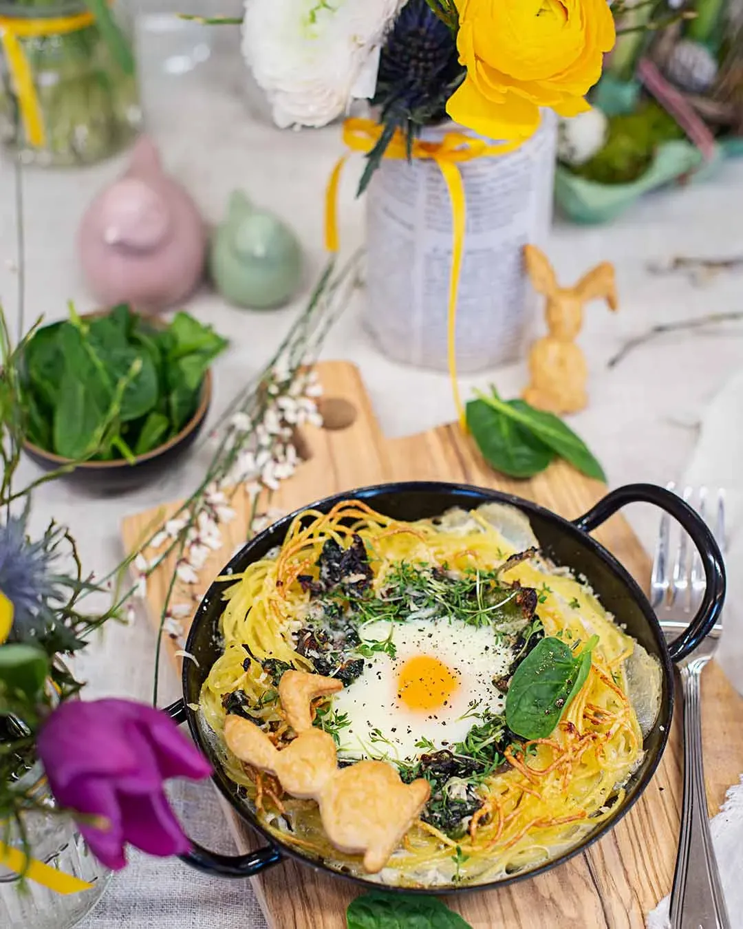 Spaghettinester für Ostern mit Spiegelei und Kräutern in einer Pfanne auf Holzbrett, mit Blumen dekoriert