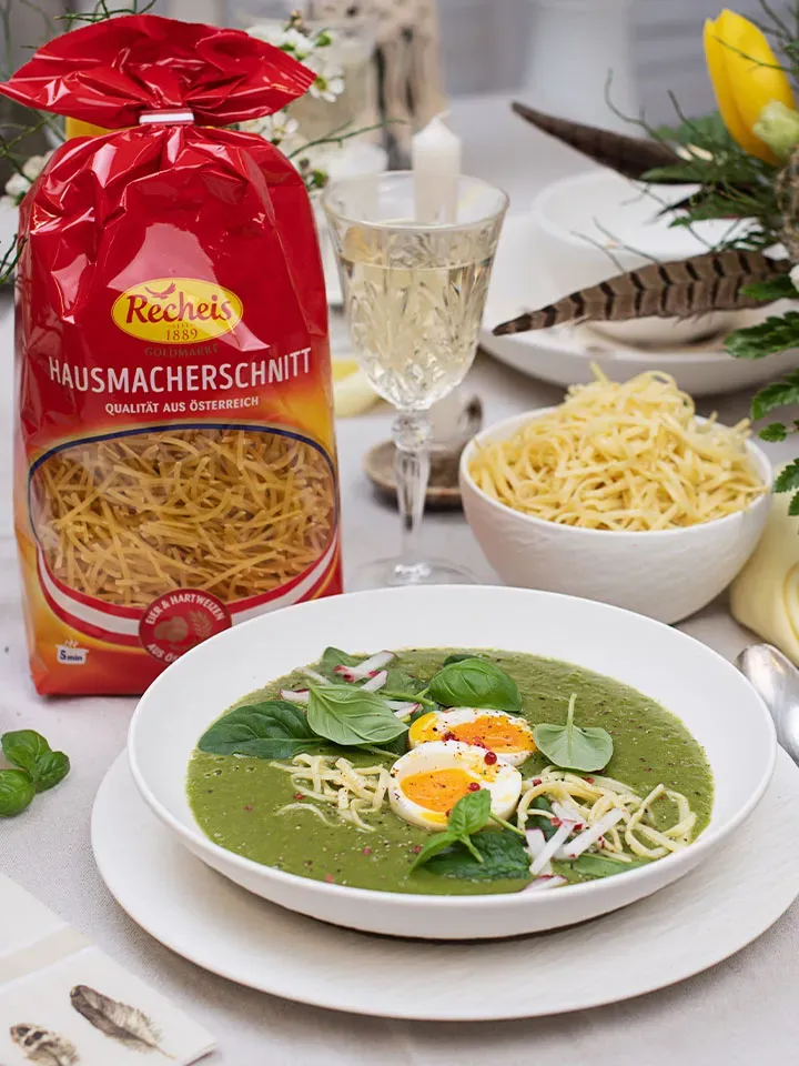 Grüne Spinatsuppe mit Ei, Nudeln und frischen Kräutern neben Nudeln und einem Glas Weißwein