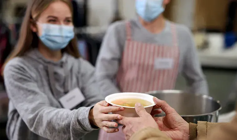 Helferin mit Mund-Nasen-Schutz reicht in einer Suppenküche eine Schale Suppe über die Theke