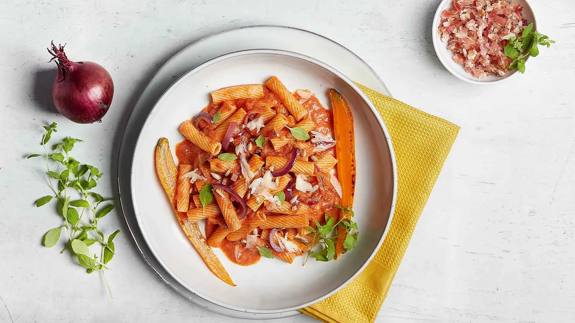 Teller mit Pasta in Tomatensauce, Zwiebeln und Kräutern auf gelbem Tuch