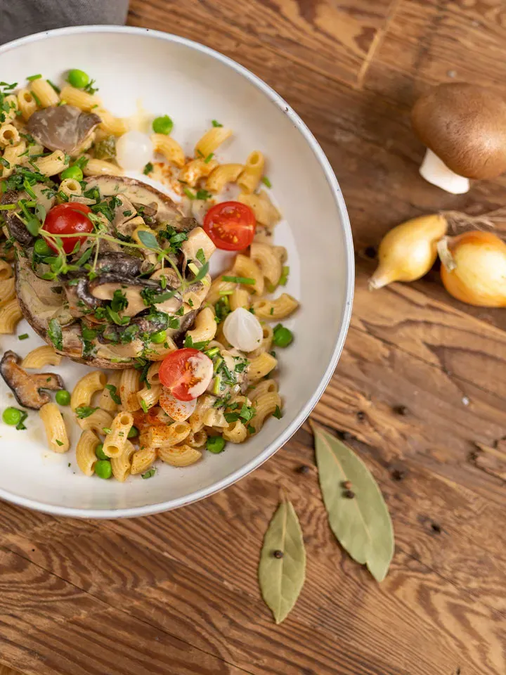Veganes Stroganoff mit Nudeln, Pilzen, Kirschtomaten und frischen Kräutern auf einem Teller auf Holztisch