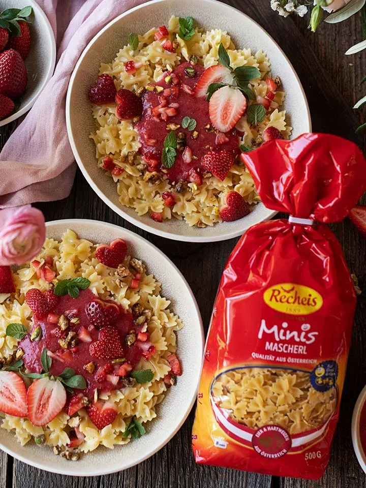 Süße Erdbeernudeln mit Mini-Mascherl und Erdbeersauce, mit frischen Erdbeeren und Minzblättern auf zwei Tellern