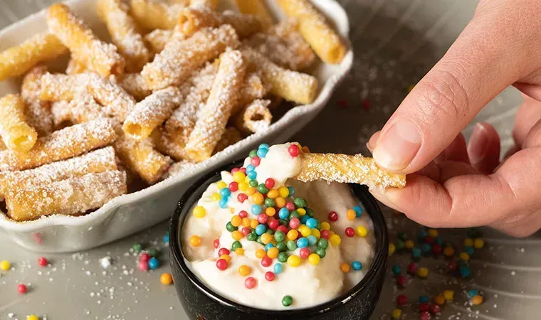 Eine Hand taucht einen Nudelchip in die Bananen-Topfen-Creme mit bunten Streuseln