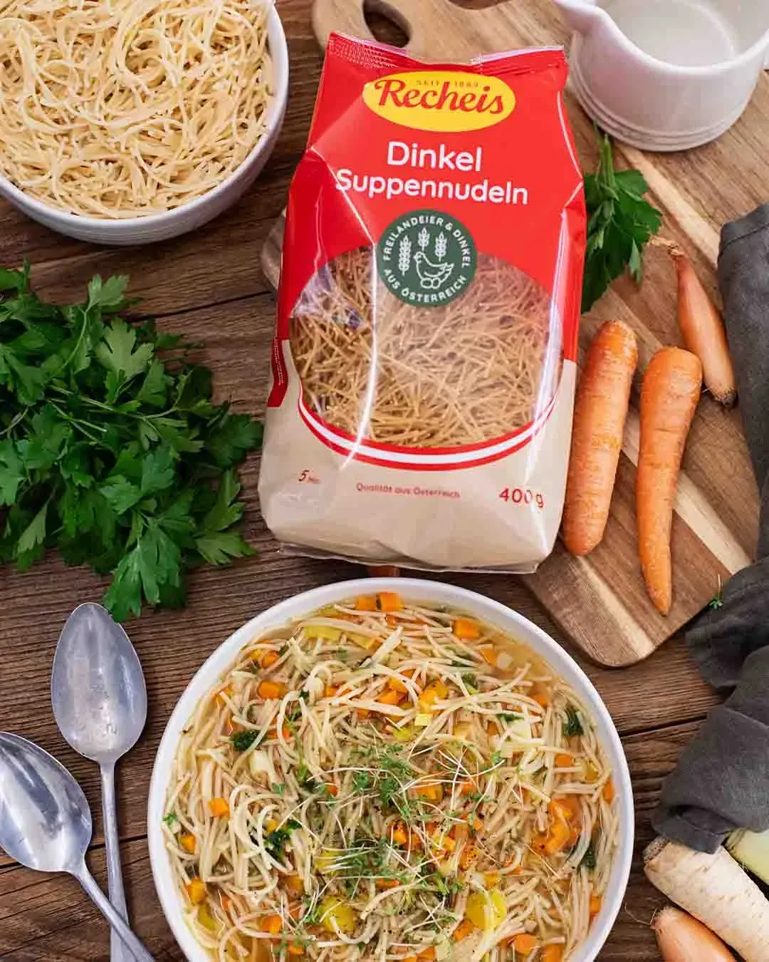 Packung Recheis Dinkel Suppennudeln neben einem Teller Gemüsesuppe mit Petersilie und Karotten auf einem Holztisch, von oben