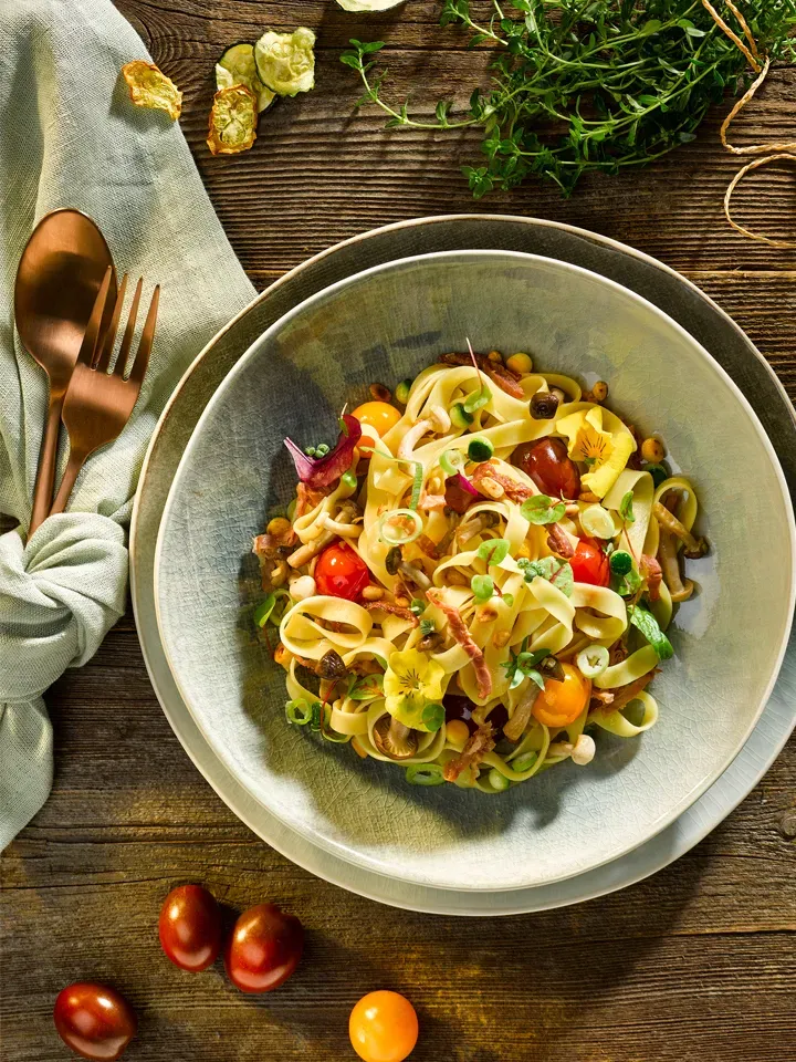 Tagliatelle Fiori mit Buchenpilzen, Zucchini, Parmaschinken, Kirschtomaten und Kräutern auf Holztisch