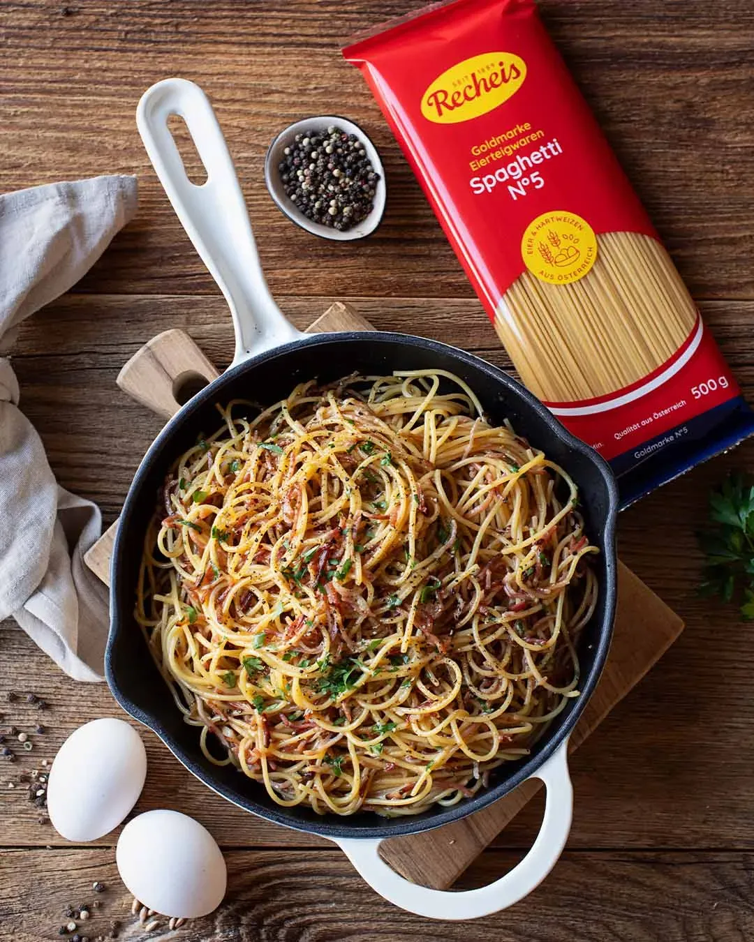 Pfanne mit Tiroler Spaghetti Carbonara, daneben Spaghettipackung, Eier und Pfefferkörner