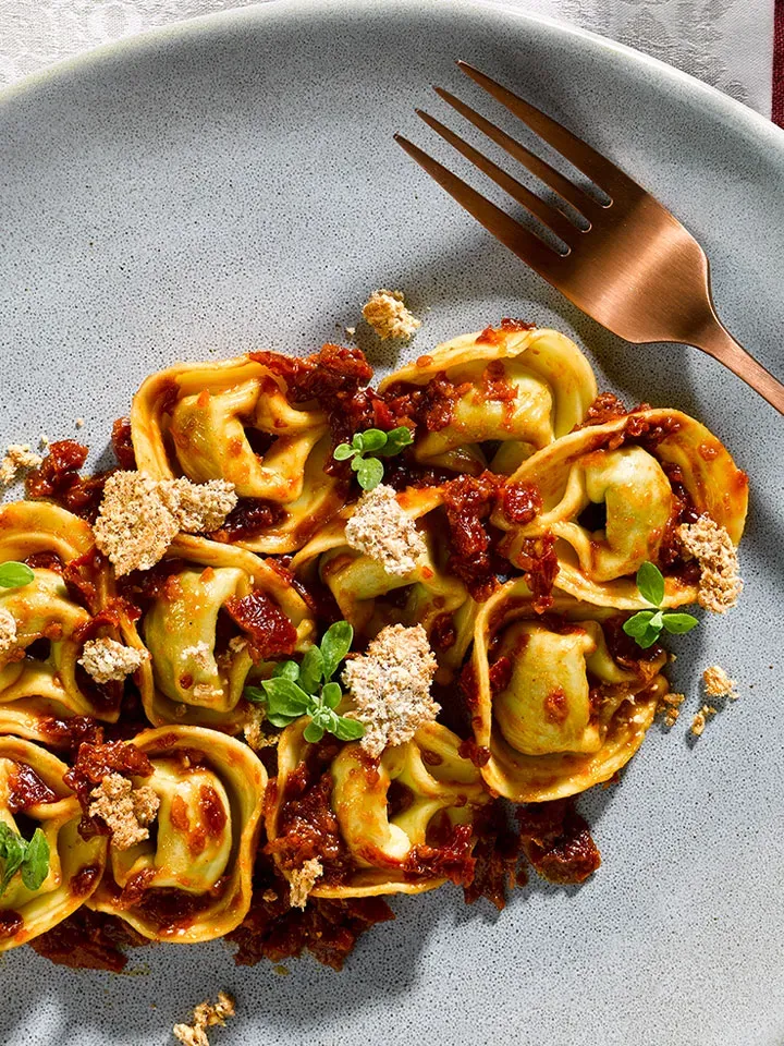 Tortelloni Spinat-Ricotta auf Tomaten-Erdnuss-Pesto, mit Kräutern und Brotkrumen, daneben eine Gabel