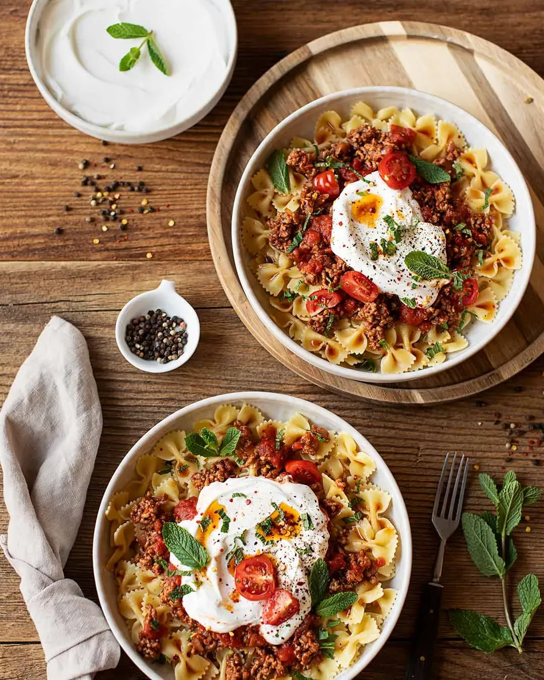 Türkische Nudeln mit Farfalle, Hackfleischsoße, Joghurt und frischen Kräutern auf zwei Tellern auf Holz