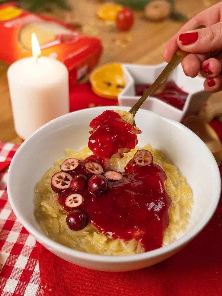 Hand gibt mit einem Löffel rote Sauce über Vanillemilchreis mit Nudeln und Cranberries in einer weißen Schüssel