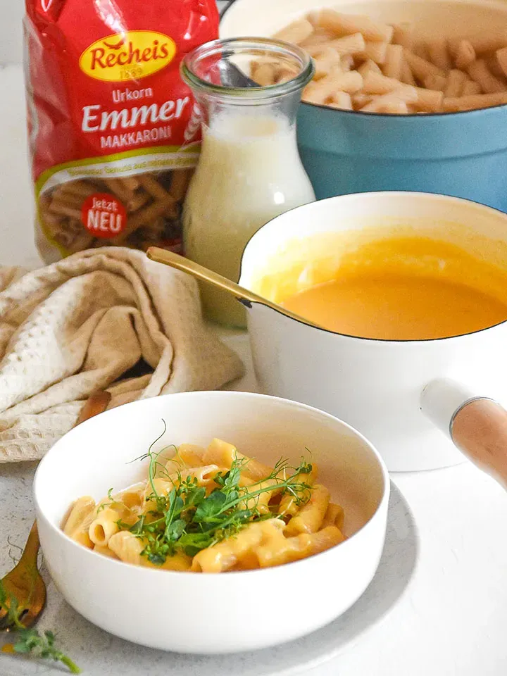 Vegane Mac and Cheese in einer Schale, im Hintergrund ein Topf mit Käsesauce und eine Milchflasche