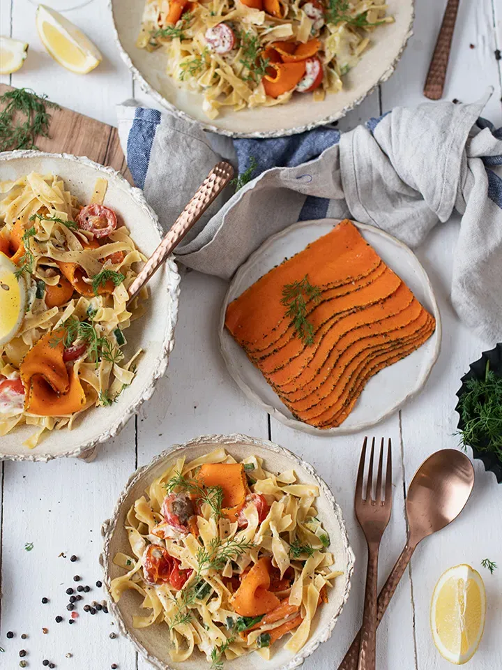 Vegane Lachsnudeln mit Vollkornbandnudeln in drei Schalen, dazu eine Platte mit dünnen Karottenscheiben auf weißem Tisch