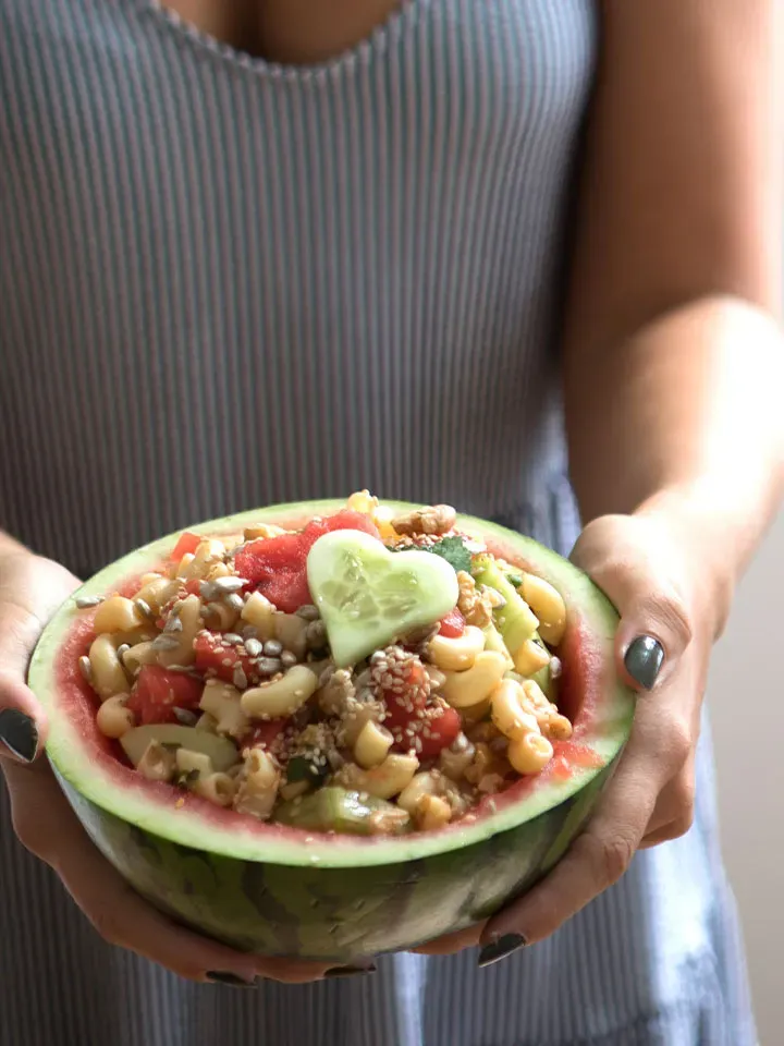 Person hält eine Wassermelonenhälfte, gefüllt mit veganem Nudelsalat und Gurkenherz