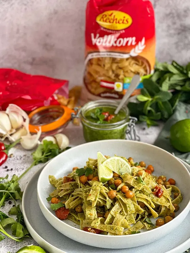 Vollkorn-Bandnudeln mit veganem Pesto, Kichererbsen und Limettenscheiben auf einem Teller, daneben Zutaten