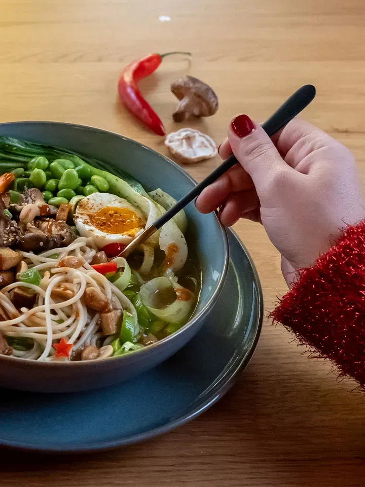 Hand mit Stäbchen über einer Schüssel wärmender Ramen mit Spaghetti, Ei, Gemüse und Chili