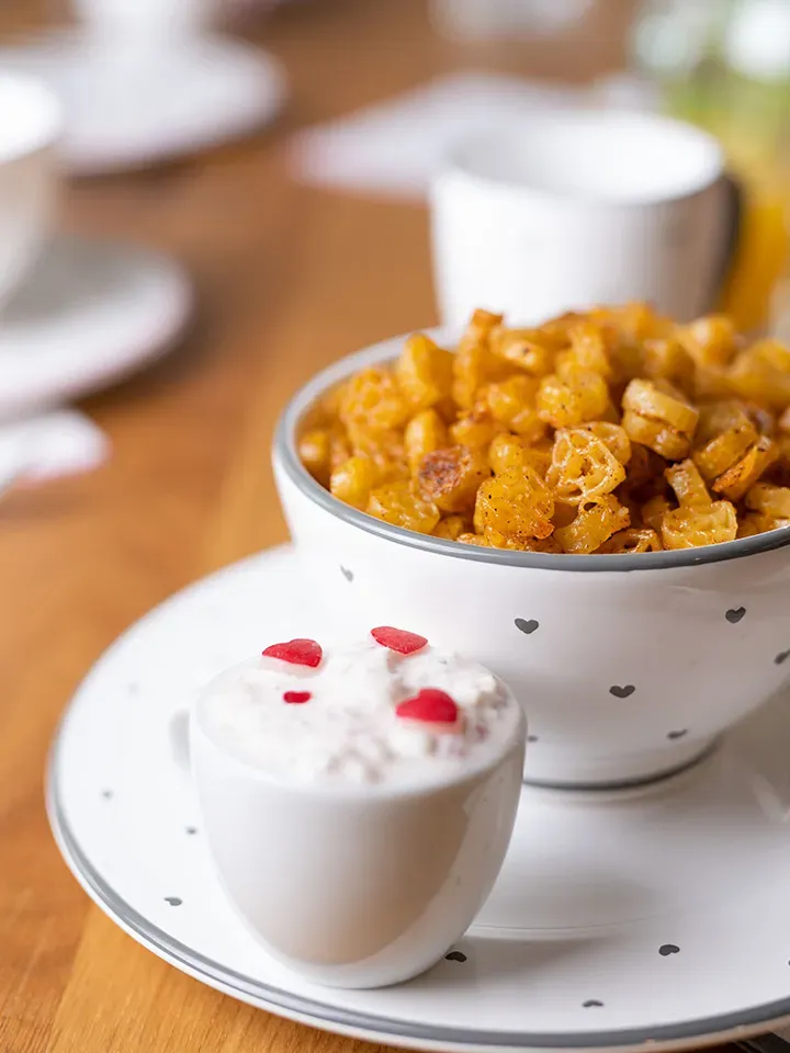 Würzige Herz-Nudelchips in einer Schüssel, daneben Tasse mit weißer Sauce und roten Stücken