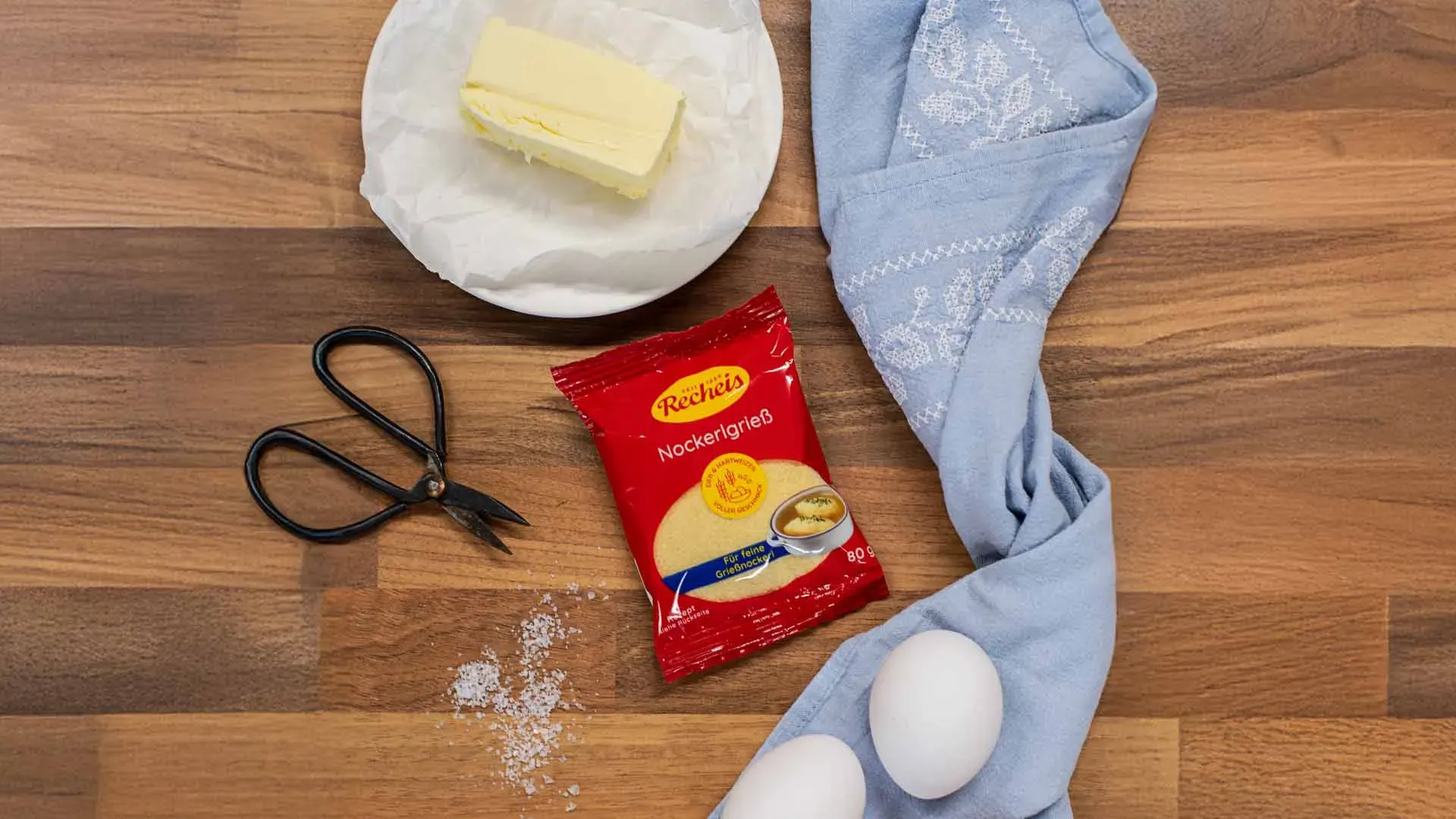 Zutaten für Grießnockerl: Butter, Eier, Grieß, Schere und blaue Stoffserviette auf Holztisch